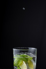 Top view of water drop falling into glass cup with lemon and mint, on black background in vertical, with copy space