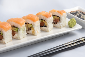 Sushi roll with pink salmon wrap, grilled salmon, Philadelphia cheese and avocado. White container and black chopsticks.