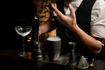 Fototapeta na wymiar close-up male bartender pours drink from metal jigger into smoky steel cup of shaker