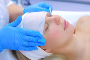 Naklejka premium Cosmetologist is wiping woman's face using antibacterial napkins, side view. Doctor is making beauty procedure in cosmetology clinic. Care about patient's face skin. Beautician cleaning girl's face.
