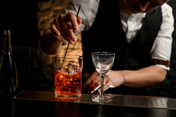 Close-up. Bartender's hand holds long spoon and mix drink in glassy mixing cup with pieces of ice