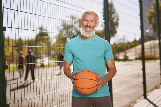 Basketball player. Happy mature man in sportswear holding basketball ball and smiling at camera while standing at outdoor basketball court