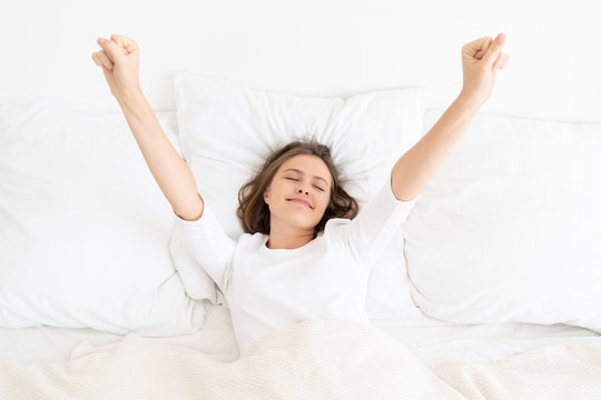 Young girl waking up in morning in white bed and pyjama top, leaving eyes closed but stretching arms up to get ready for active day, showing sleepy smile