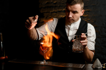 Close-up. Man at bar gently sprinkles on glass with cocktail and make fire.