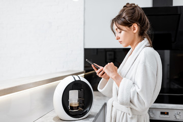 Young woman in bathrobe using a smart phone while making coffee with a coffee machine on the...