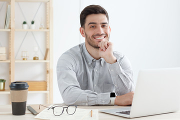 Business guy wearing gray shirt, sitting in front of open laptop, looking at camera with positive smile, ready to help and answer questions