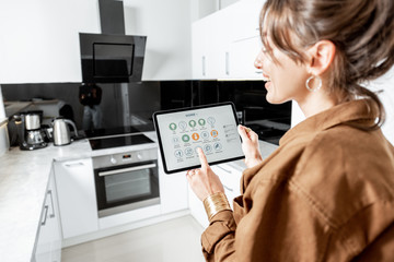 Young woman controlling kitchen appliances with a digital tablet, mobile device with launched smart...