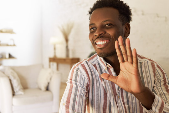 Body language and positive human emotions. Picture of joyful friendly young dark skinned man sitting in stylish apartment interior greeting friend with hand, looking with happy broad smile