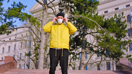 Young man in a yellow jacket and hat wearing his protective mask while standing on the park stairs new Covid-19