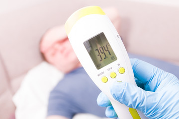 hand in a rubber glove and a medical gown measure the temperature of a man on an infrared non-contact thermometer, high temperature 39.4 degrees Celsius, a man on the sofa blurred background