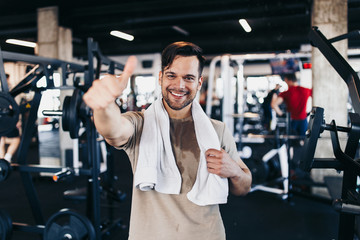 Young handsome man after successful workout posing and smiling in modern fitness gym while showing thumb up