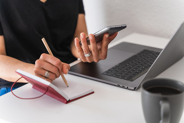 Woman working while writing notes in a notebook with mobile phone in hand and a laptop in front of her