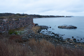 The rocky shore and the ancient granite wall of the fortification of Suomenlinna Island and a view of the bay and islands at autumn twilight in Finland. The harsh and laconic beauty of Finnish nature.
