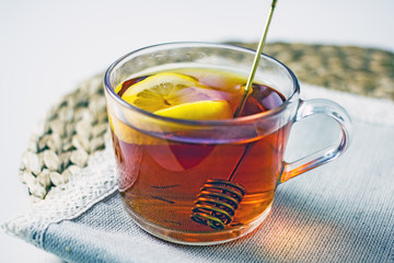 A transparent mug with black tea, a slice of lemon, a spoon for honey on a wicker gray napkin on a light table. The concept of a cozy tea party. Close-up