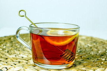 A transparent mug with black tea, a slice of lemon, a spoon for honey on a wicker gray napkin on a light table. The concept of a cozy tea party. Close-up