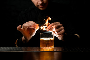 Close-up bartender gently sprinkle to alcoholic cocktail with piece of citrus zest and make fire...