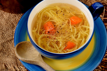 Chicken soup with noodles in a blue mug next to a wooden spoon on a rustic table