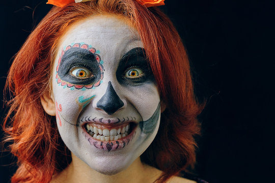 Young woman in day of the dead mask skull face art make-up and red hair showing teeth on dark background close up. Halloween concept, mad smile