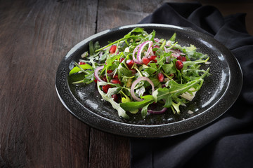Fresh rocket salad or arugula with pomegranate seeds, red onions and feta cheese on a black spotted plate on a dark wooden table, copy space
