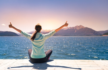 Happy young woman feeling energized, with thumbs up sitting by a beautiful mountain lake at sunrise. 