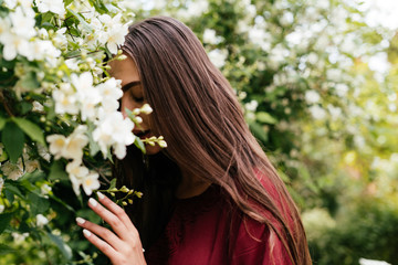 Naklejka premium romantic long-haired brunette in a cherry-colored dress buried her face in jasmine flowers and gently covering her eyes enjoys the aroma