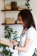 indoor portrait of young beautiful woman standing and reading book. She is wearing black wrist watch, bookshelf is in the background.