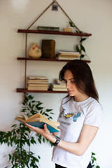 indoor portrait of young beautiful woman standing and reading book. She is wearing black wrist watch, bookshelf is in the background.