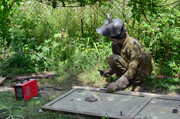 Old man welder in brown uniform, welding mask and welders leathers, weld metal door with arc...
