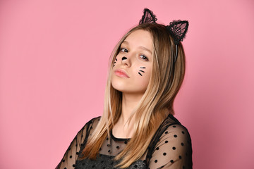 Teenage girl in black dress, headband like cat ears, bracelet. Looking at you, hands folded, posing on pink background. Close up
