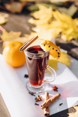 Transparent tall glass with red mulled wine boiled from grape wine on wooden background with autumn leaves, oranges and spices