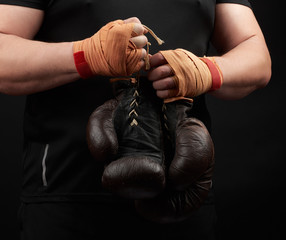 athlete in a black uniform holds very old brown boxing gloves in his hand