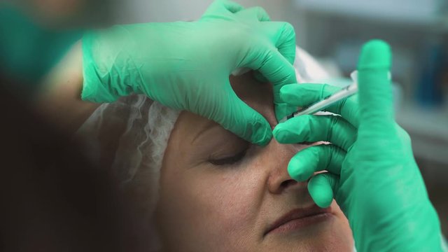 CU: skilled specialist in color gloves does anti-aging dysport injections into middle aged woman nose bridge with syringe in beauty salon closeup