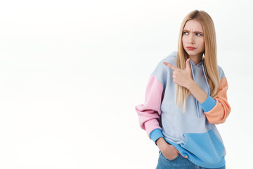 Portrait of doubtful, suspicious blonde girlfriend having hesitation, squinting and frownign skeptical, looking pointing finger left with judgement and disappointment, dont like product