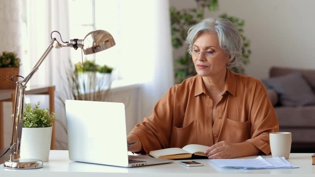  Elderly lady in casual clothes using laptop and making notes then reading document while working on remote business project at home
