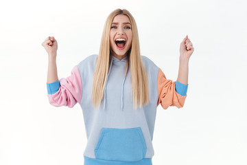 Happy, excited blonde girl in hoodie, chanting, celebrating victory, winning prize, achieve goal, raising hands up in hooray, smiling broadly, triumphing over white background