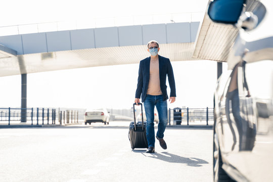 Outdoor shot of adult man returns from business trip, goes to car with suitcase, wears medical mask during coronavirus outbreak, pandemic disease. Passenger arrives after interantional traveling.