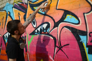 Young graffiti artist with backpack and gas mask on his neck paints colorful graffiti in pink tones on brick wall. Street art and contemporary painting process