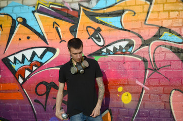 Young caucasian graffiti artist in black t-shirt with silver aerosol spray can near colorful graffiti in pink tones on brick wall. Street art and contemporary painting process