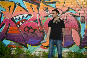 Young caucasian graffiti artist in black t-shirt with silver aerosol spray can near colorful graffiti in pink tones on brick wall. Street art and contemporary painting process
