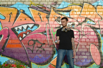 Young caucasian graffiti artist in black t-shirt with silver aerosol spray can near colorful graffiti in pink tones on brick wall. Street art and contemporary painting process