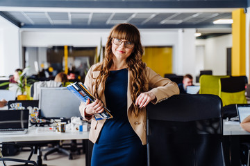 Smart girl with glasses, wearing classic office clothes, stands in coworking space, leaning her hand on a chair and looking to the camera. Other freelancers working on computers on  background