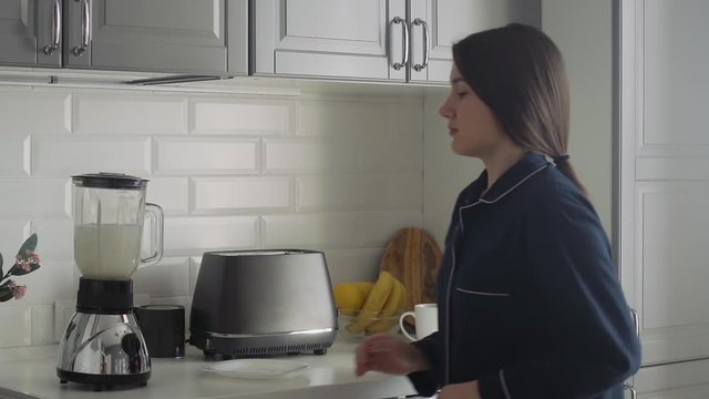 Young Woman In Black Pajamas Preparing Breakfast At The Morning. Woman Turns On Toaster, Electric Kettle And Makes A Cocktail In A Blender