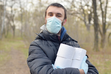 Covidiot concept. Young man in protective mask holds many rolls of toilet paper outdoors in spring...
