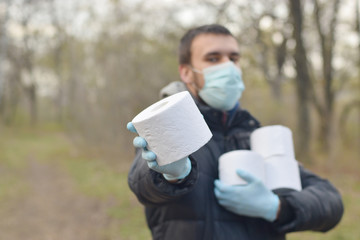 Covidiot concept. Young man in protective mask holds many rolls of toilet paper outdoors in spring...