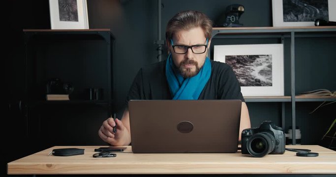 Bearded photographer in eyeglasses, back t-shirt and blue scarf retouching photos on laptop in studio. Concept of professional equipment and creativity