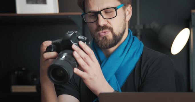 Cheerful bearded man in black t-shirt and blue scarf reviewing photos on camera. Professional photographer in eyeglasses during working process in studio.