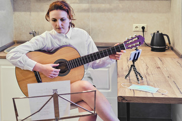 Guitarist plays a musical instrument in the home kitchen in front of the phone. Learning to play the guitar during the coronavirus pandemic.