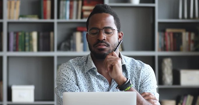 Head shot pensive young biracial man looking at laptop screen, thinking of hard problem decision or case study. Thoughtful mixed race entrepreneur businessman employee stack with difficult task.