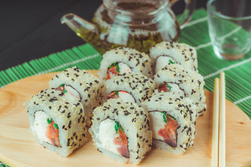 Delicious rolls on wooden board with soy sauce and green tea on decorative bamboo napkin. Close up of tasty sushi rolls with chopsticks on cutting board on black background.