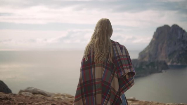 A young blonde woman walks on the cliff to the edge, in the background the island of Es Vedrà and the cloudy sky, full hd medium shot slow motion video